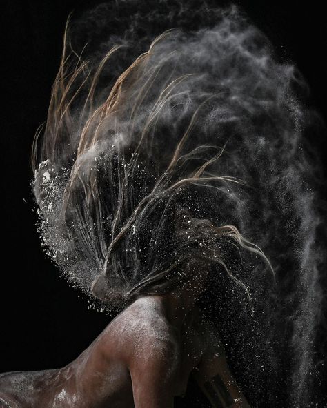 Artistic image of a person shaking their head with a dramatic spray of powder or dust flowing through their hair, captured against a dark background. The motion and particles create a dynamic, ethereal effect, blending movement and stillness