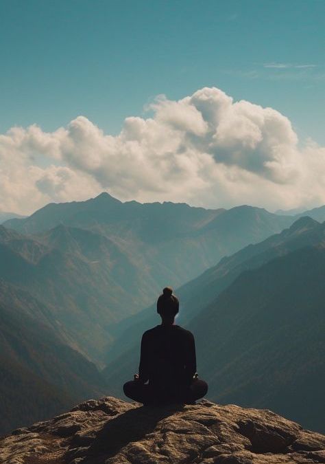 A person sits atop a mountain, gazing at the expansive view of surrounding peaks and valleys in the distance.